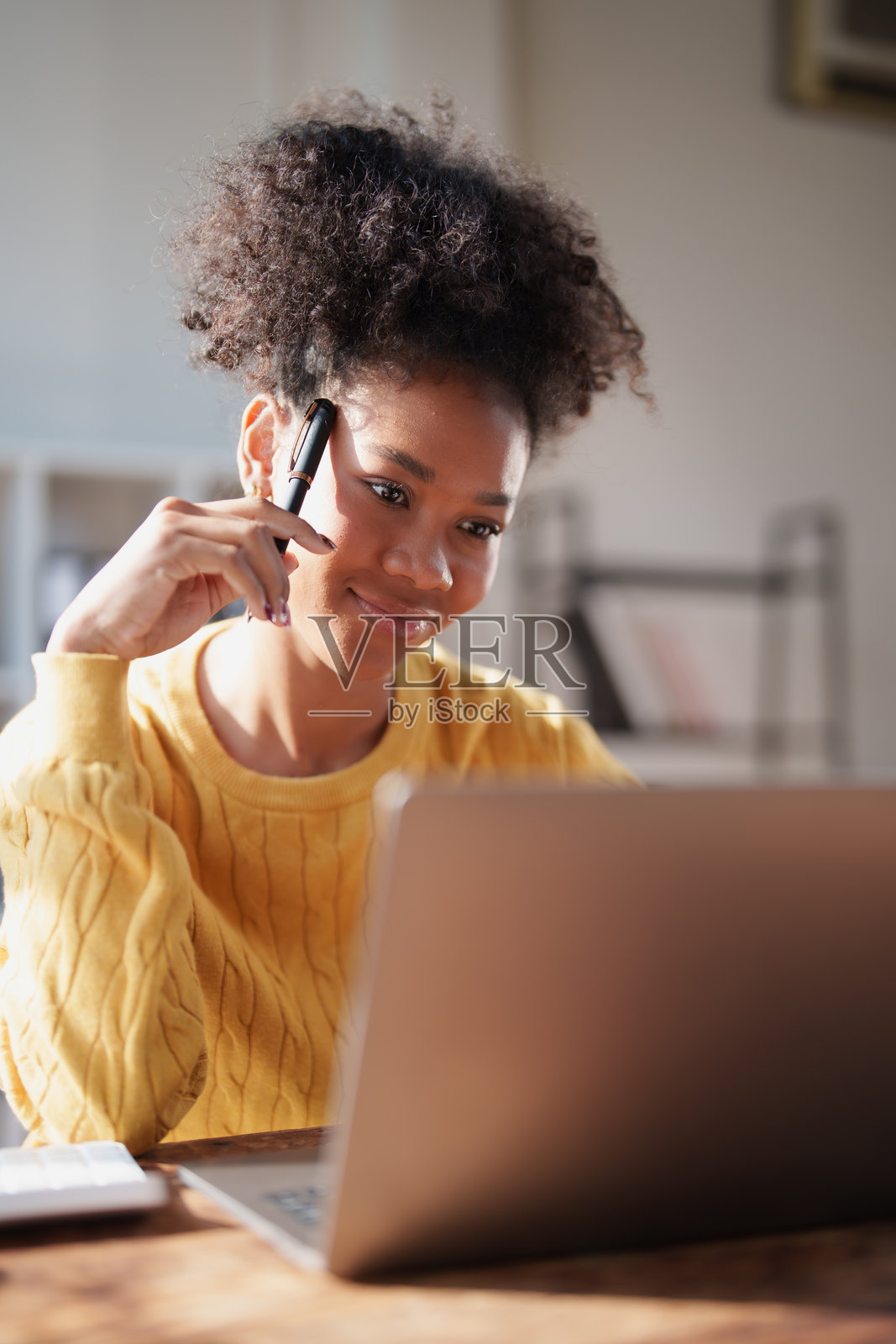 一位年轻女性在家中明亮的办公室环境中，手持钢笔，目光注视着笔记本电脑屏幕，积极思考并在线协作，体现了远程工作与学习。照片摄影图片