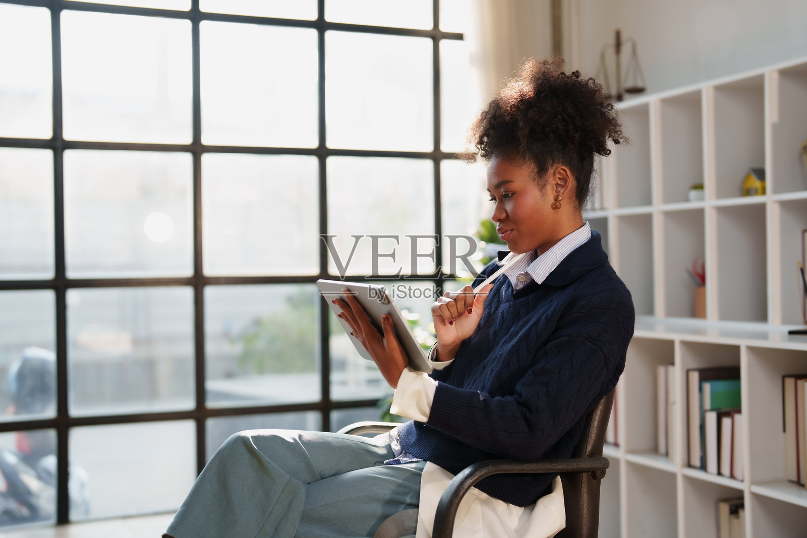 一位年轻的黑人女性坐在一间明亮现代的办公室里，她手持一支触控笔的数码平板电脑，在浏览在线内容和处理专业任务时面带微笑。照片摄影图片