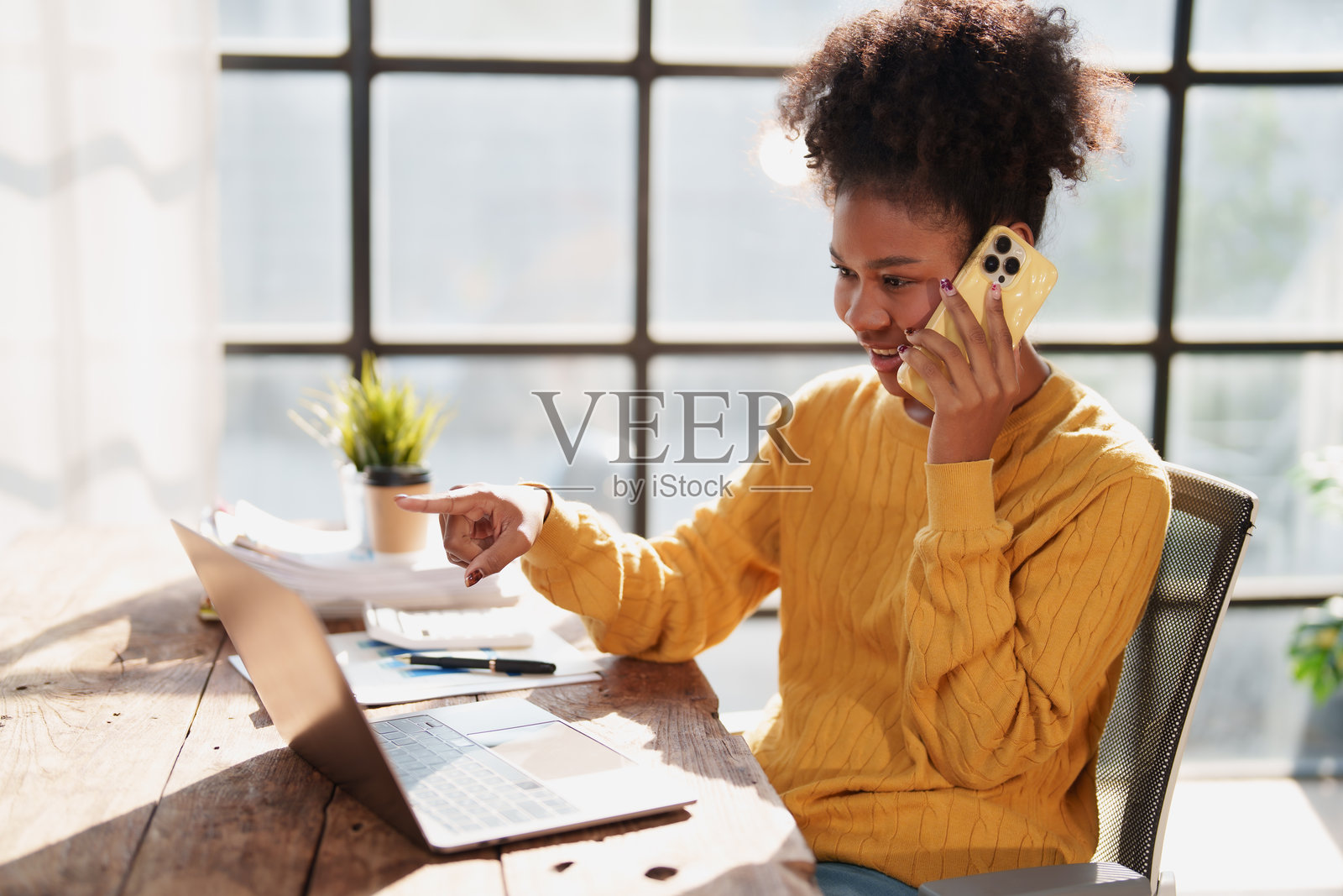 年轻女子在家远程办公，一边用智能手机打电话，一边用手指着笔记本电脑屏幕，强调沟通与效率照片摄影图片