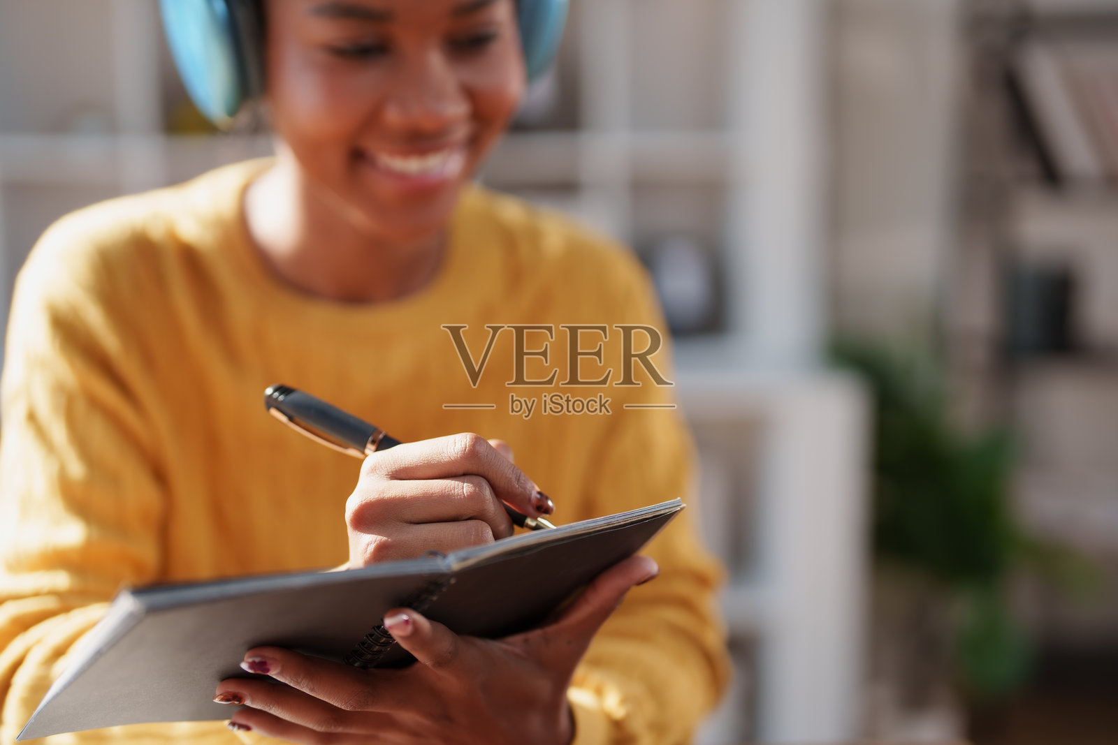 一位年轻的黑人女性，戴着蓝色耳机，穿着黄色上衣，面带微笑，正用笔在小笔记本上书写，在明亮的室内环境中记录想法或学习。照片摄影图片