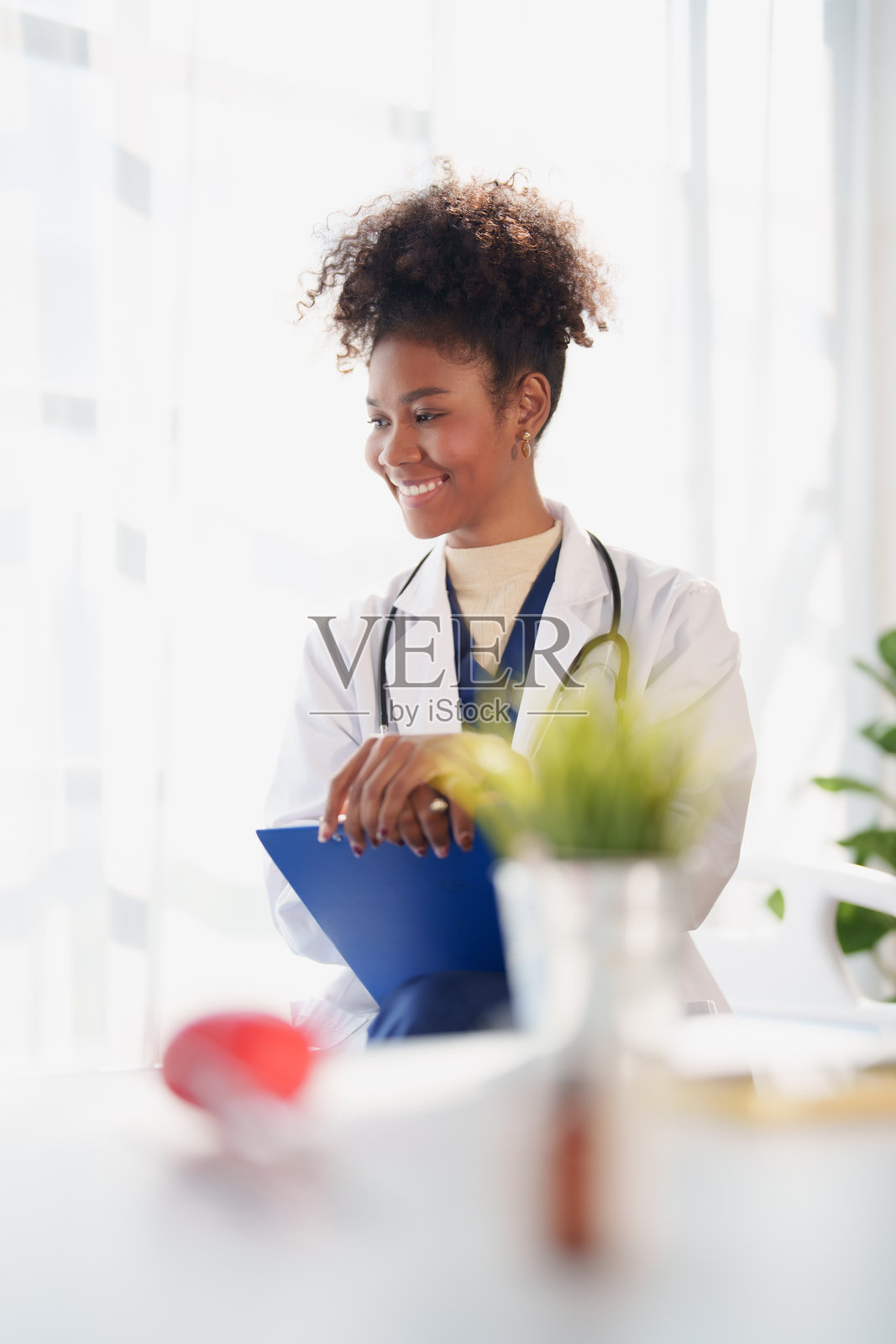 年轻女医生身穿白大褂，戴着听诊器，在明亮的诊室里进行医疗咨询时，微笑着拿着一个蓝色的病历夹照片摄影图片