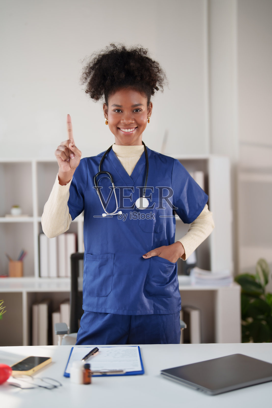 年轻的非裔美国女性医护人员，身穿蓝色手术服，戴着听诊器，面带微笑，用食指向上指着，在诊所内站立。照片摄影图片