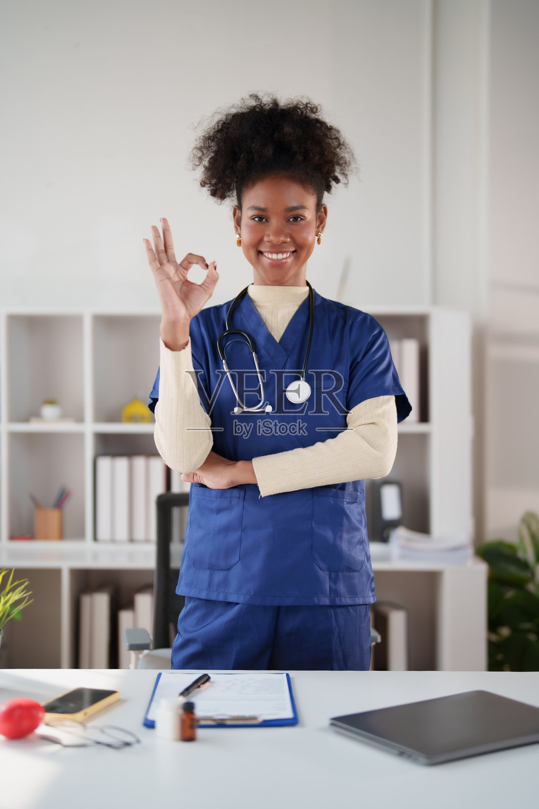 一位年轻的非裔美国女性医务工作者，身穿手术服，戴着听诊器，在诊所内自信地微笑，并竖起大拇指表示赞许和关怀。照片摄影图片