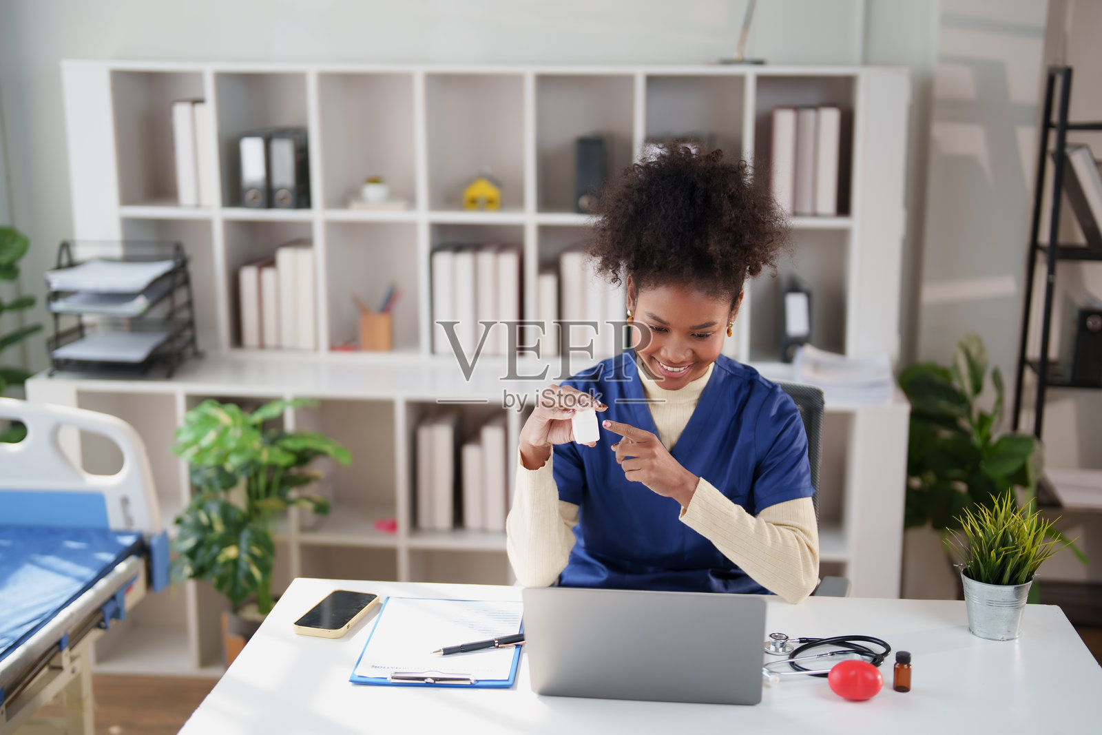 一位身穿手术服的非裔女医生，在诊室里通过笔记本电脑进行远程医疗咨询，她微笑着指向处方药瓶，向患者解释药物并进行虚拟问诊。照片摄影图片
