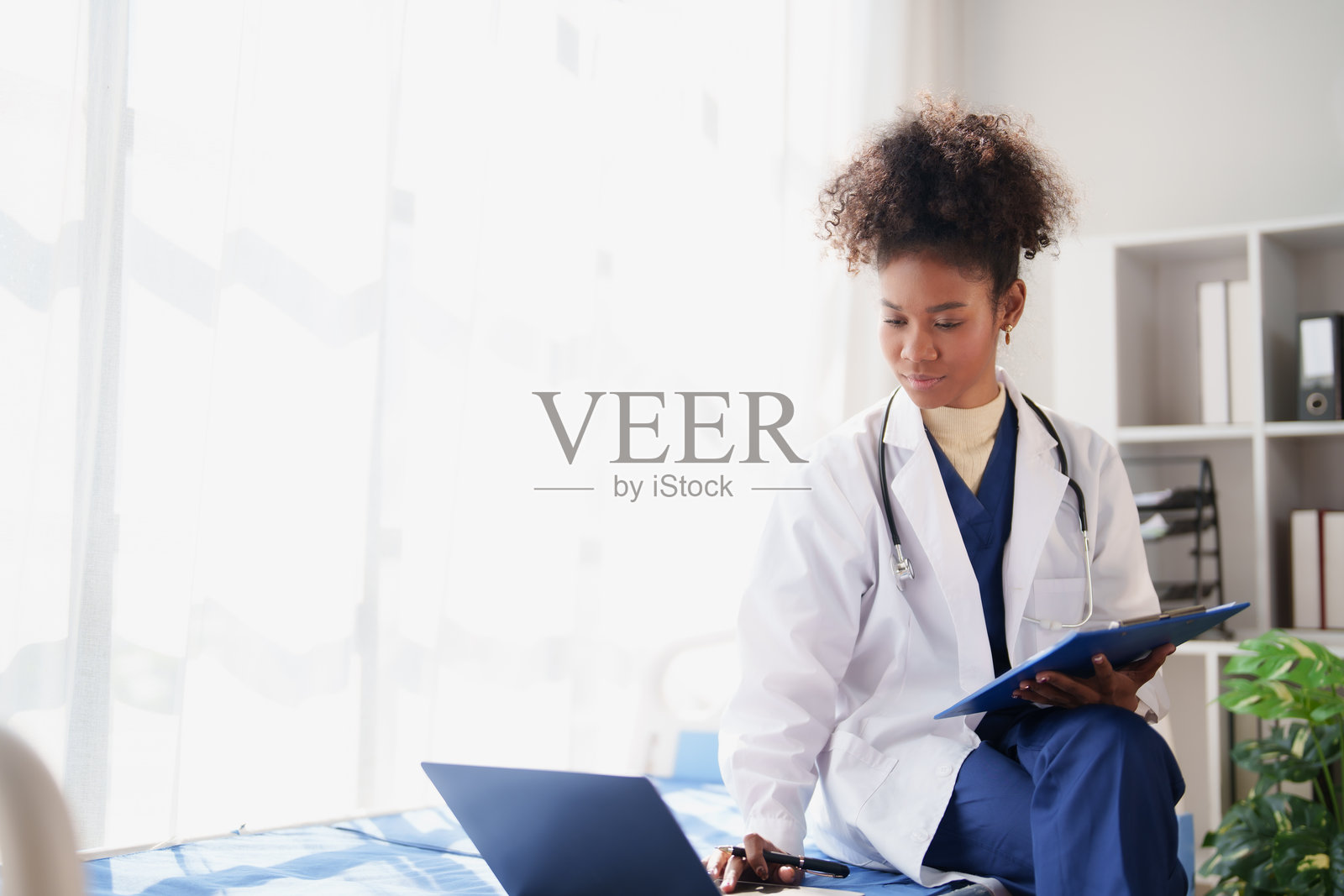 年轻的非裔美国女医生，身穿白大褂，戴着听诊器，坐在明亮的诊所病床上，正在用笔记本电脑和剪贴板查看电子病人记录。照片摄影图片