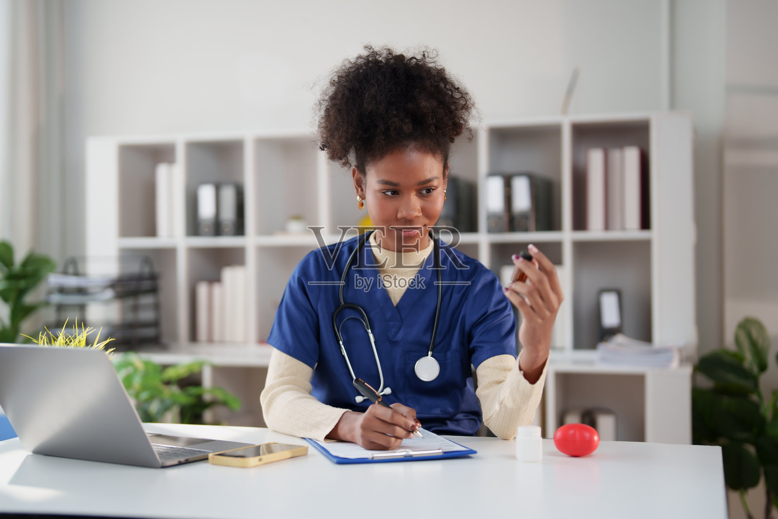 一名身穿工作服、佩戴听诊器的年轻非洲裔女护士，手持药瓶，在诊所或办公室环境中用夹板书写笔记。照片摄影图片