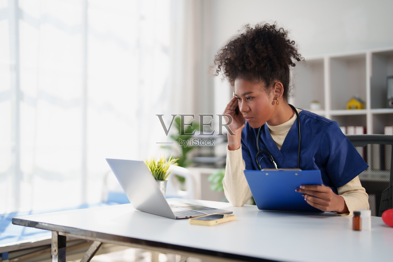 身穿蓝色工作服、佩戴听诊器的女医生正在办公室里专注地操作笔记本电脑，手中拿着一个写字板，画面象征着医疗保健技术和医学咨询。照片摄影图片
