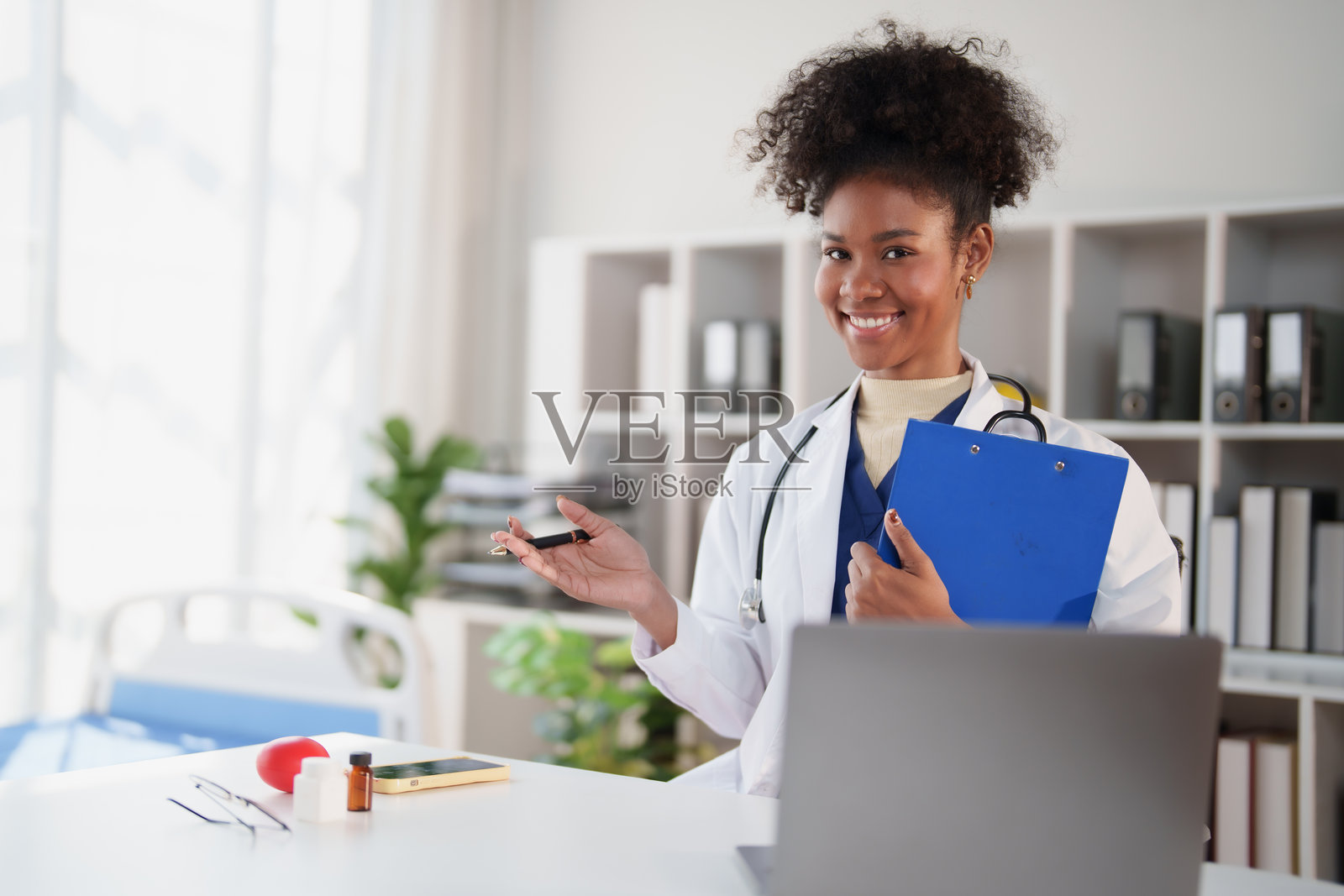 一位穿着白大褂、戴着听诊器的非裔美国女医生，坐在办公桌前，电脑上显示着远程会议，她微笑着，一边拿着剪贴板和笔一边做着手势。照片摄影图片
