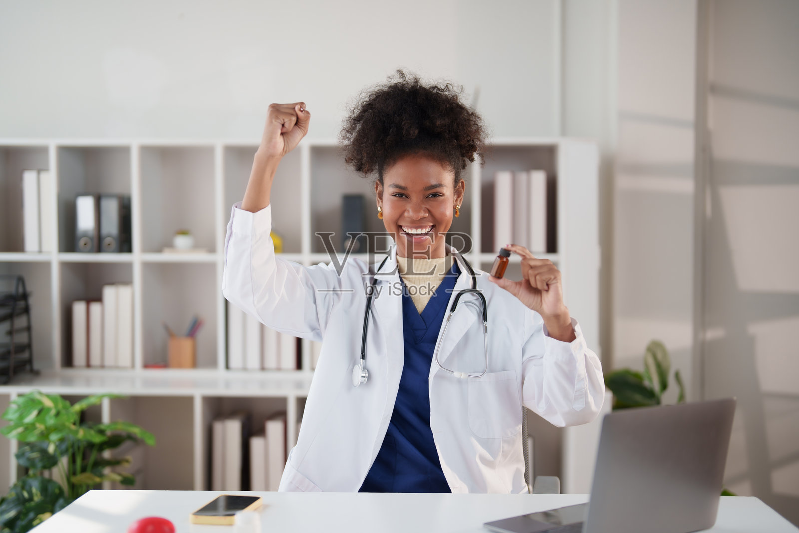 年轻的女性医生，身穿白大褂，戴着听诊器，面带微笑，高举拳头，庆祝医学突破，手中还拿着一小瓶棕色药剂。照片摄影图片