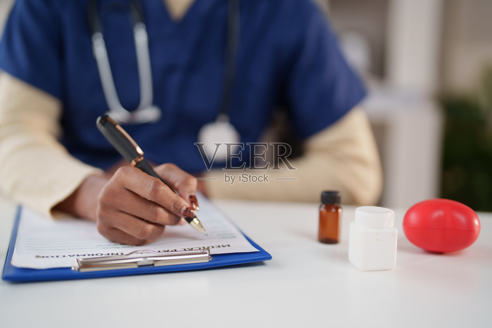 一名身穿手术服、佩戴听诊器的医疗专业人员正在夹板上记录患者的医疗详细信息，桌上放有药瓶和心脏压力球。照片摄影图片