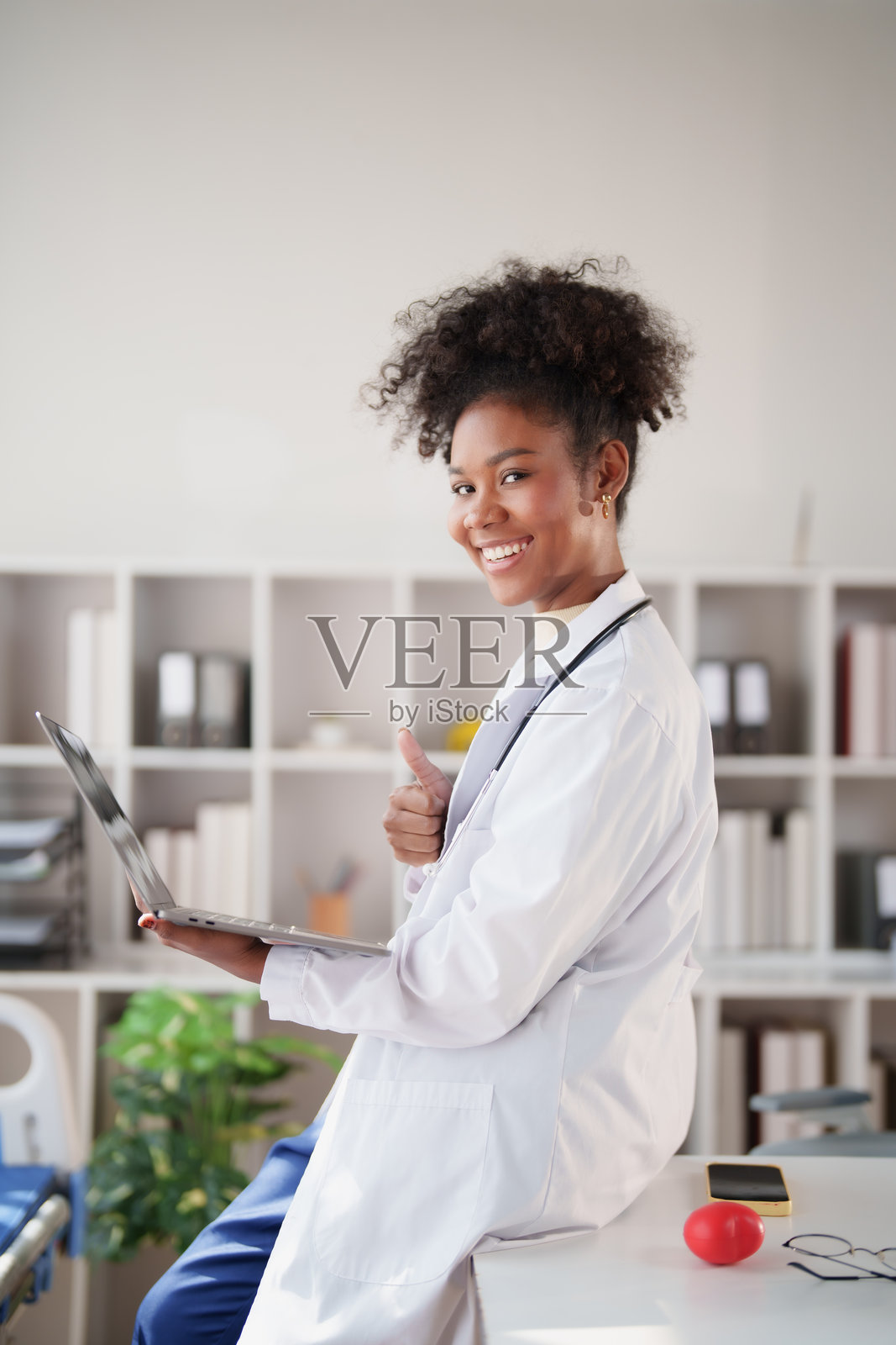 年轻的非裔美国女医生，身穿白大褂，戴着听诊器，微笑着竖起大拇指，在现代诊所里自信地准备进行远程医疗咨询。照片摄影图片