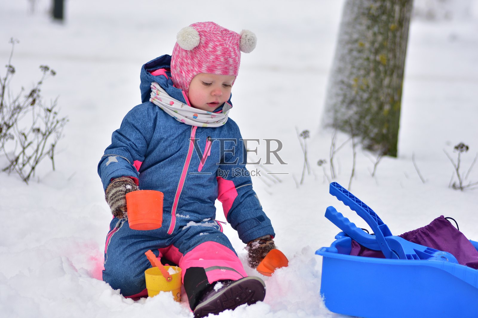 一名幼儿在户外玩雪，手持彩色塑料杯和雪橇。孩子穿着温暖的冬季雪地服，享受雪天、创意玩耍、冬季活动和童年乐趣。照片摄影图片