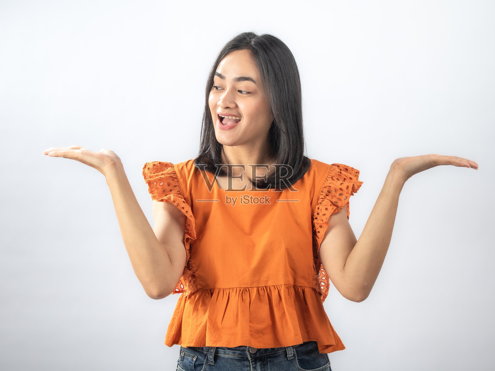 一位东南亚女性，黑色直发，身穿橙色荷叶边衬衫和牛仔裤，脸上带着惊喜而愉悦的表情，双手举在脸颊两侧，背景为纯白色。照片摄影图片