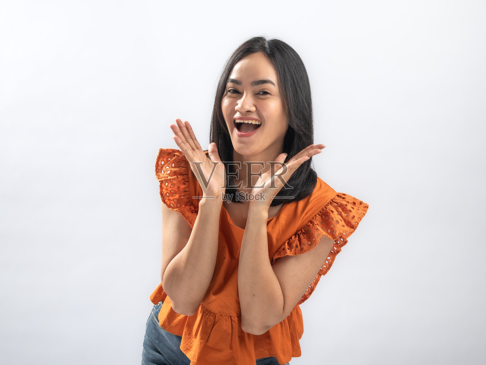 一位面带灿烂笑容的东南亚女性，留着黑色直发，身穿橙色褶皱上衣和牛仔裤，双手俏皮地举在脸颊旁，背景是纯白色。照片摄影图片