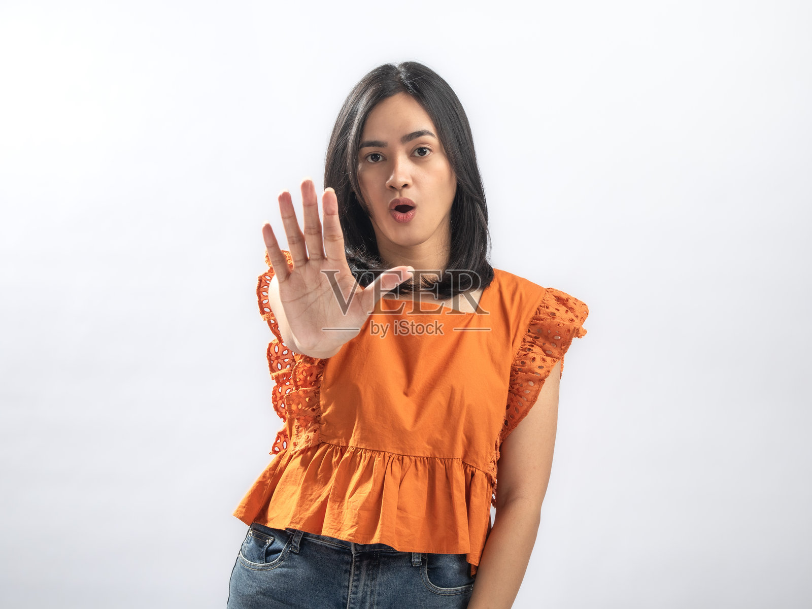 一位东南亚女性，留着黑色直发，身穿橘色荷叶边衬衫和牛仔裤，双手掌心向前，做出停止的手势，表情严肃，背景为纯白色。照片摄影图片