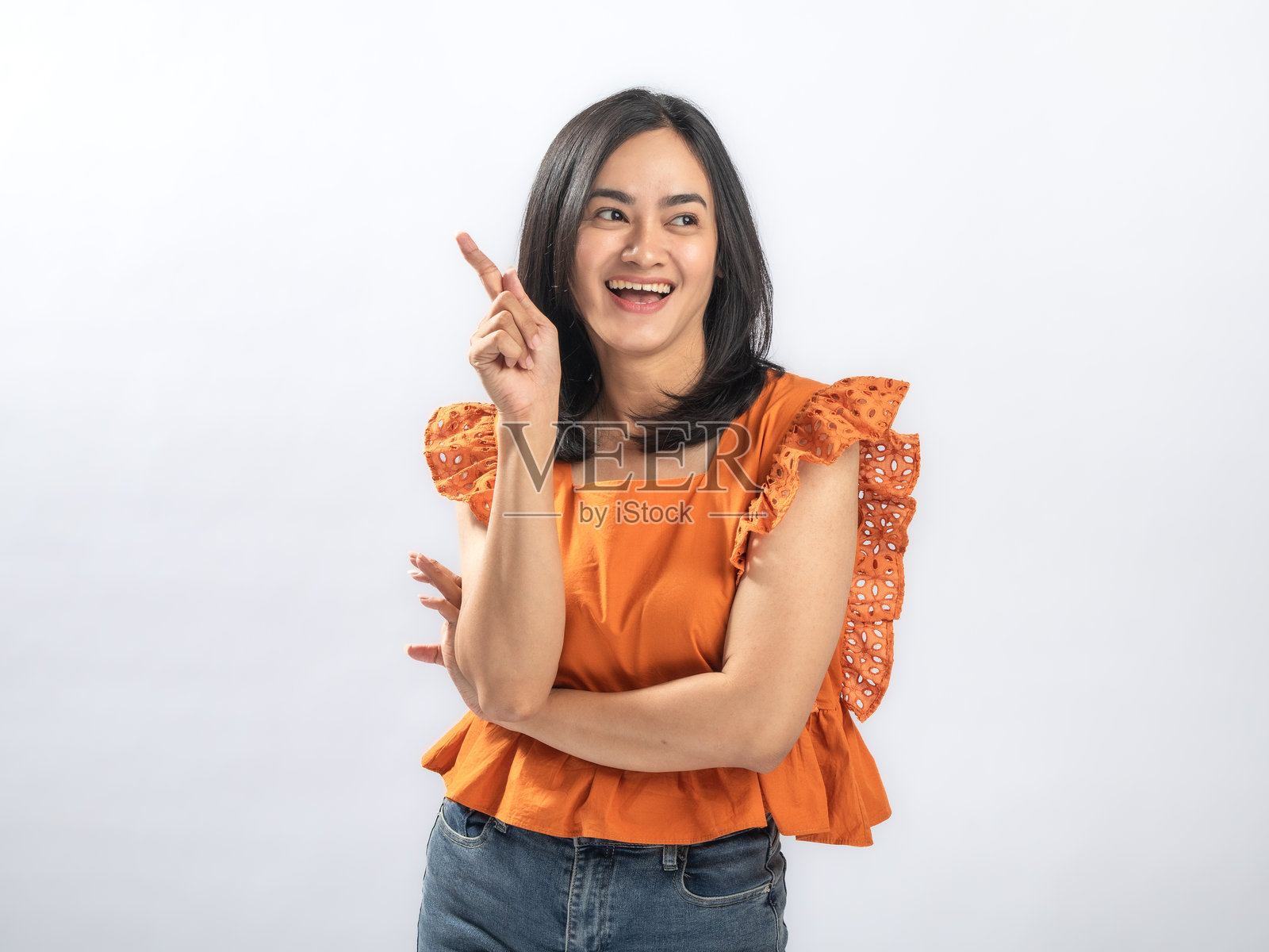 一位兴奋的东南亚女性，黑直发，笑容灿烂，身穿橙色荷叶边上衣和牛仔裤，食指向上指向，站在纯白色背景前照片摄影图片