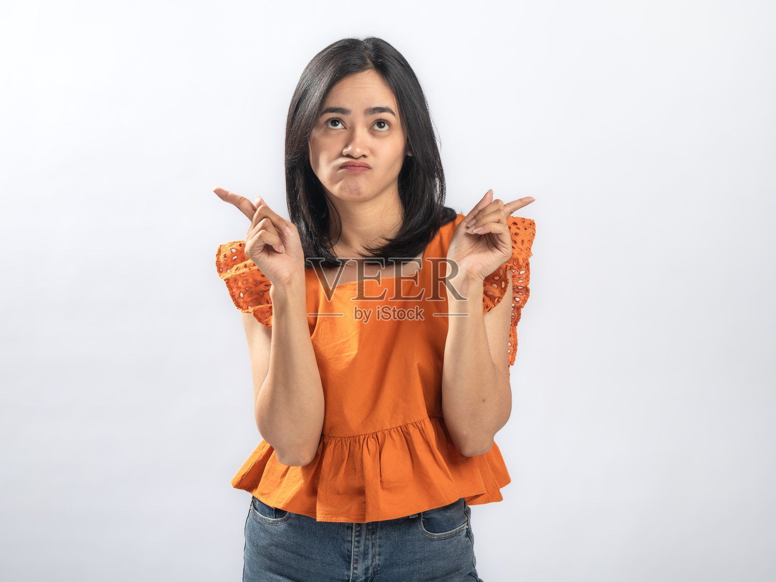 一位兴奋的东南亚女性，黑直发，笑容灿烂，身穿橙色荷叶边衬衫和牛仔裤，双手食指向上竖起，站在纯白色背景前照片摄影图片