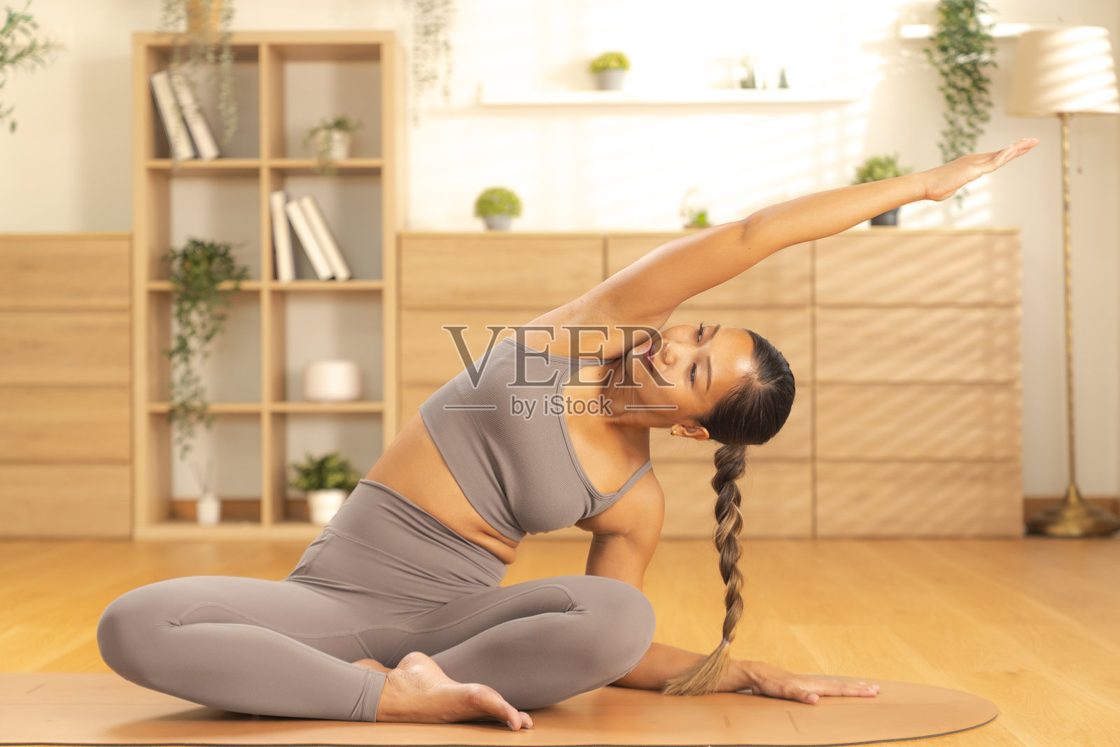 身着灰色运动服的年轻女子坐在瑜伽垫上，一条手臂向上伸展，正在做侧身拉伸，表情平静专注，头发编成辫子，背景是明亮简约的客厅照片摄影图片