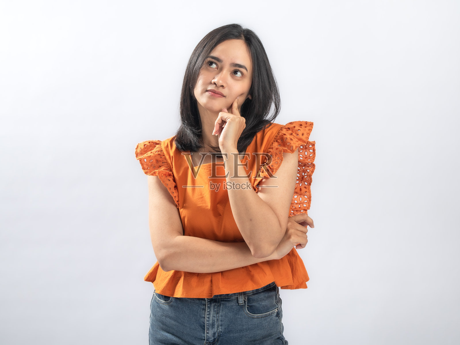 一位沉思的东南亚女性，留着黑色直发，穿着橙色荷叶边衬衫和牛仔裤，站在白色背景前，用食指触碰脸颊，若有所思地向上看。照片摄影图片
