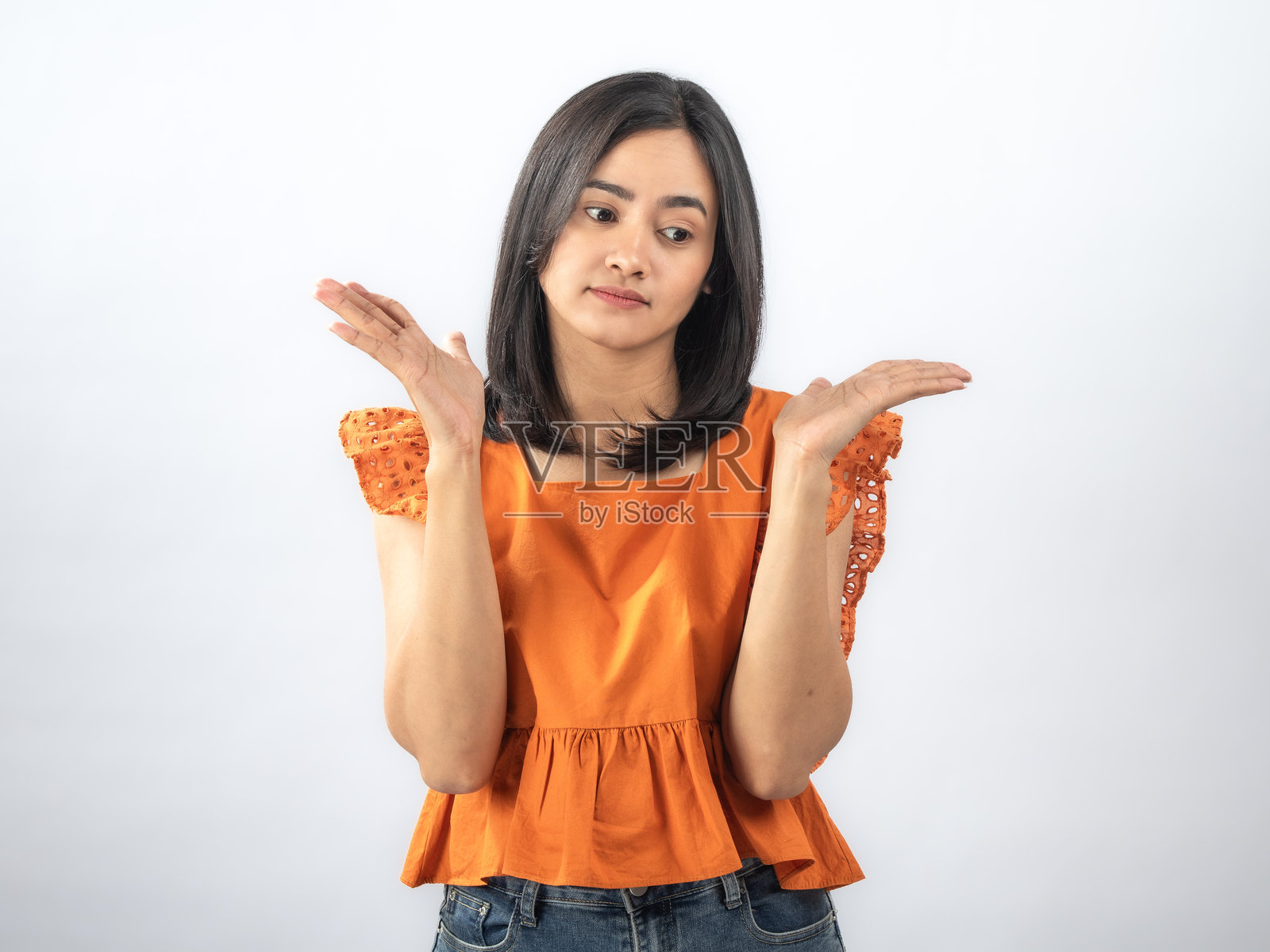 一位东南亚女性，黑直发，穿着橙色荷叶边衬衫和牛仔裤，脸上带着惊喜的表情，双手举在脸颊两侧，背景为纯白色照片摄影图片