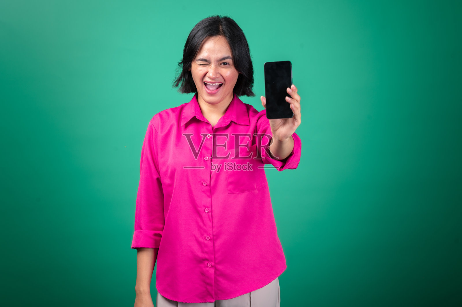 一位穿着亮粉色衬衫的亚洲女性，双臂交叉，对着绿色背景露出灿烂的笑容，同时举着一部智能手机。照片摄影图片