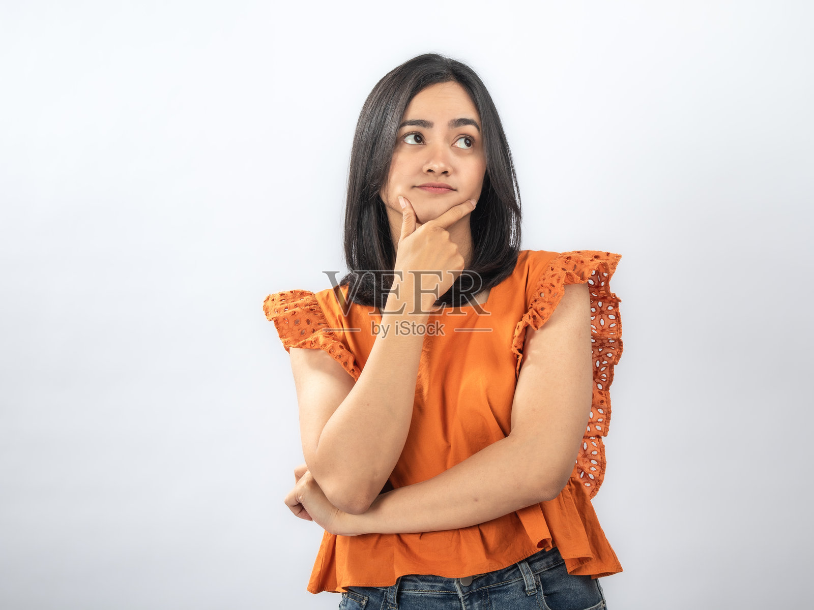 一位沉思的东南亚女性，留着黑色直发，身穿橙色荷叶边衬衫和牛仔裤，手托下巴，若有所思地站着，背景是纯白色照片摄影图片