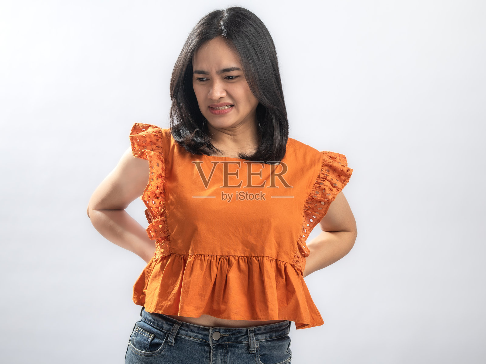 一位留着长黑发的亚洲女子，身穿橙色荷叶边衬衫和牛仔裤，一手扶着腰部，痛苦地龇牙咧嘴，表情显示腰痛或肌肉拉伤，背景为纯白色照片摄影图片