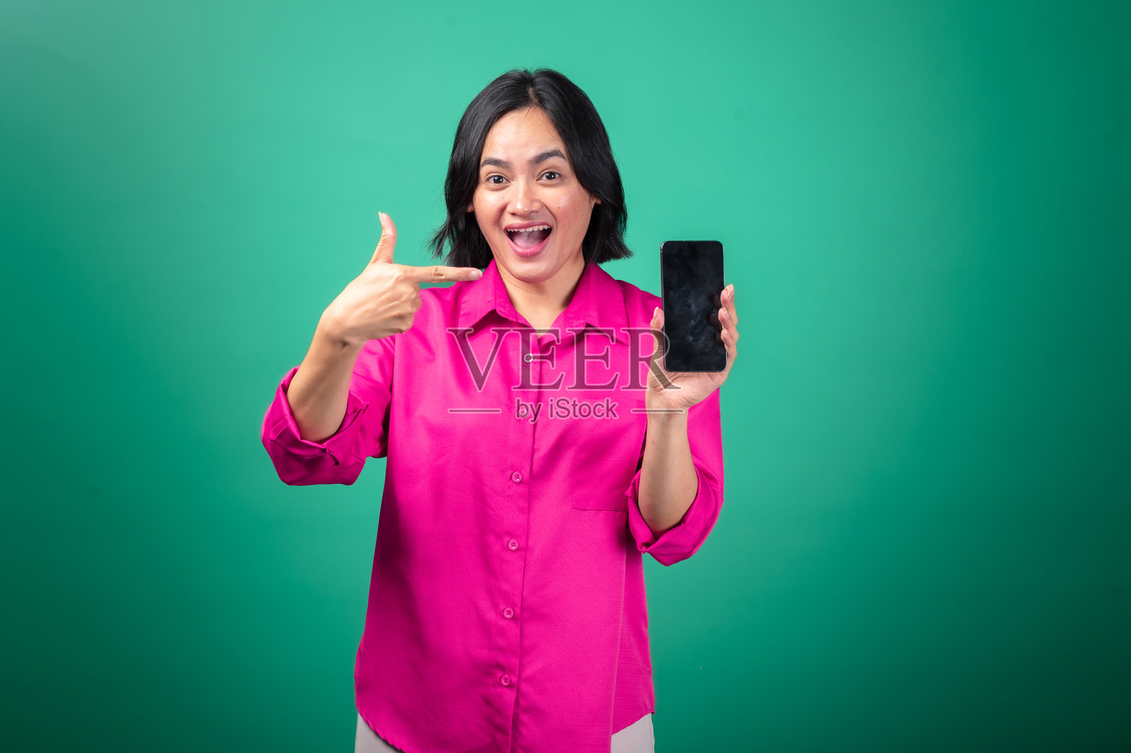 一位穿着亮粉色衬衫的亚洲女性，脸上带着灿烂的笑容，兴奋地举着一部智能手机，指向屏幕，背景为绿色。照片摄影图片
