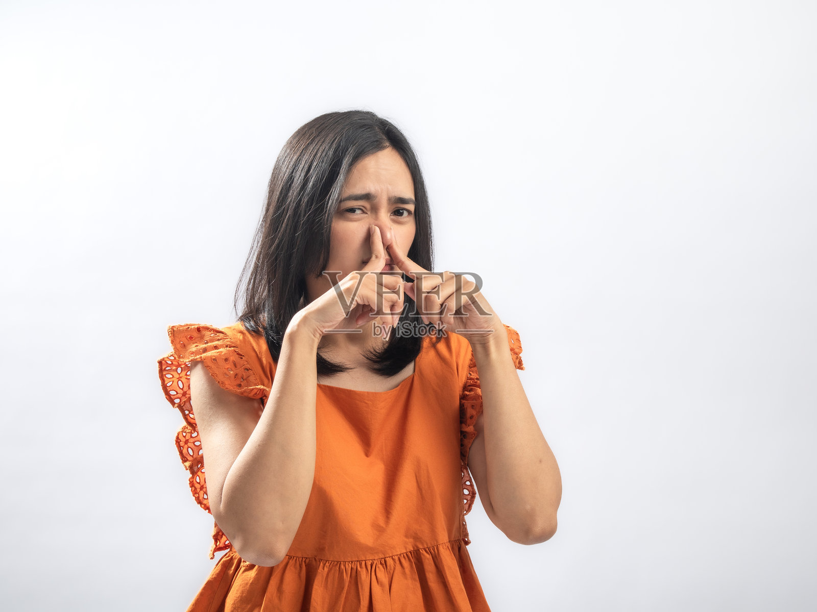 一位亚洲女子穿着橙色褶皱衬衫，捏着鼻子，面露厌恶的表情，对难闻的气味做出反应，她站在纯白背景前。照片摄影图片