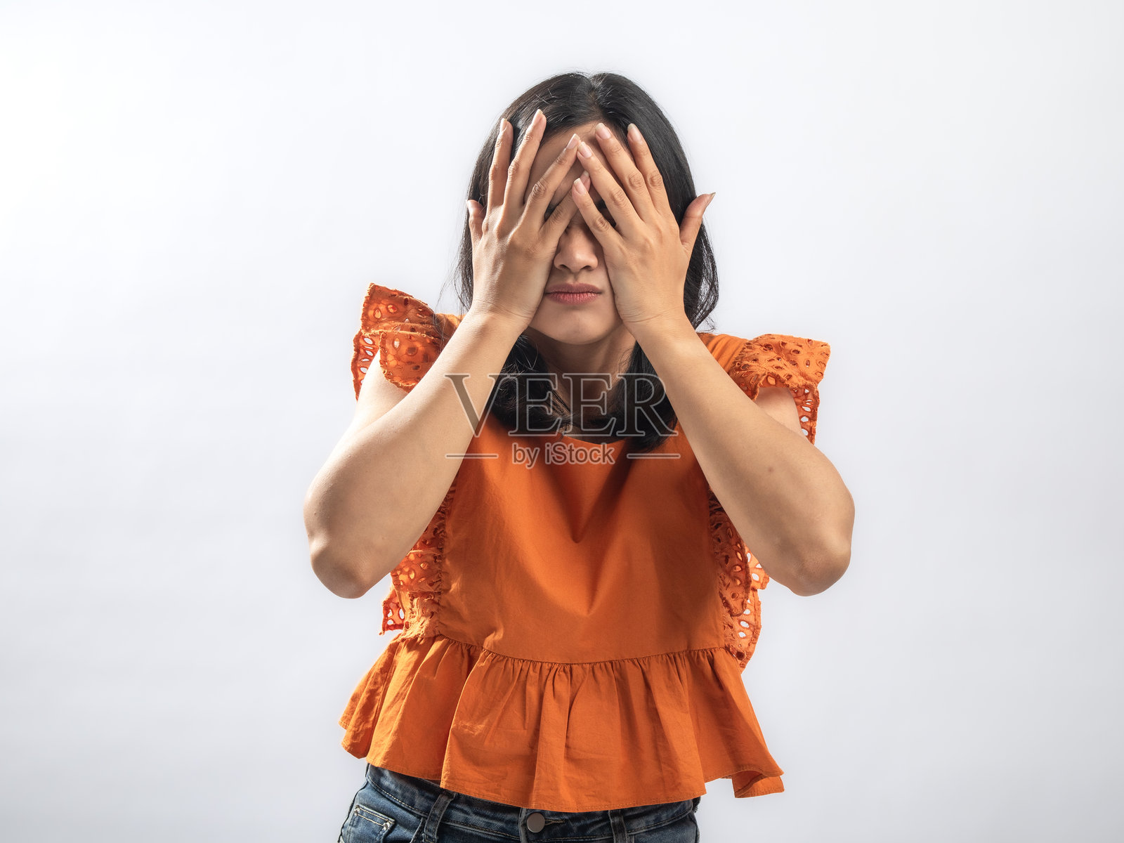 一位亚洲女性身穿橙色荷叶边衬衫，双手遮脸，表情沮丧地透过手指缝隙偷看，在纯白色背景的摄影棚内拍摄情绪肖像照照片摄影图片