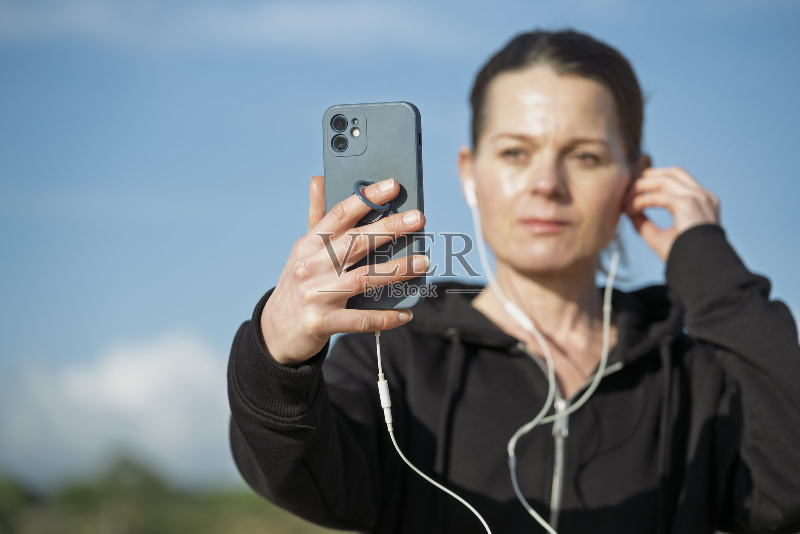 身穿黑色连帽衫的运动风女子正用智能手机佩戴白色耳机自拍。照片摄影图片