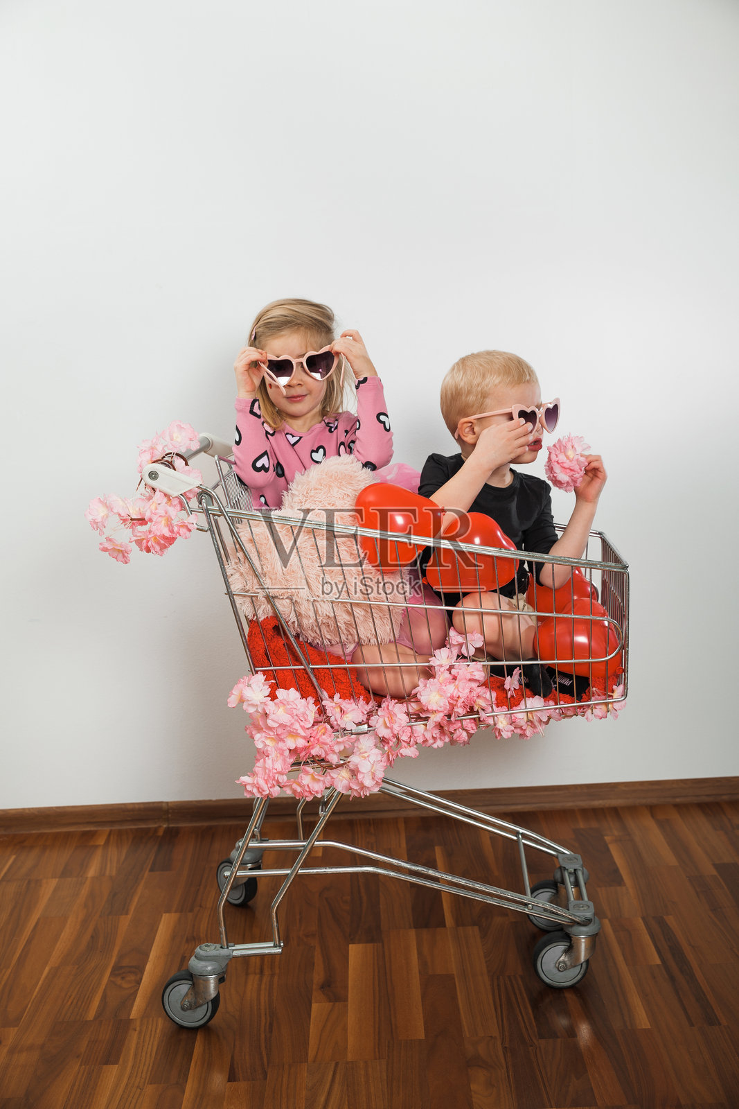 两个孩子坐在购物车里，购物车里装着气球和玩具，身处一间室内房间照片摄影图片
