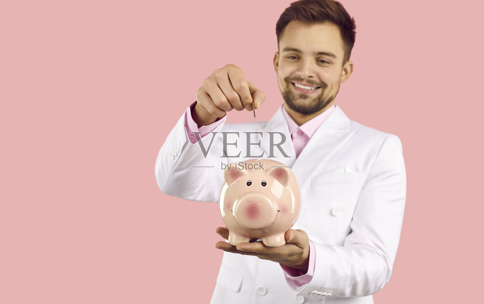 快乐的年轻商人将硬币放入他的存钱罐，孤立在粉红色背景上照片摄影图片