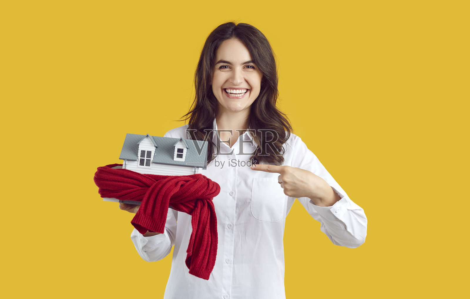 一名快乐的女子站在橙色背景前，用手指着一个用针织围巾包裹的房屋模型。照片摄影图片
