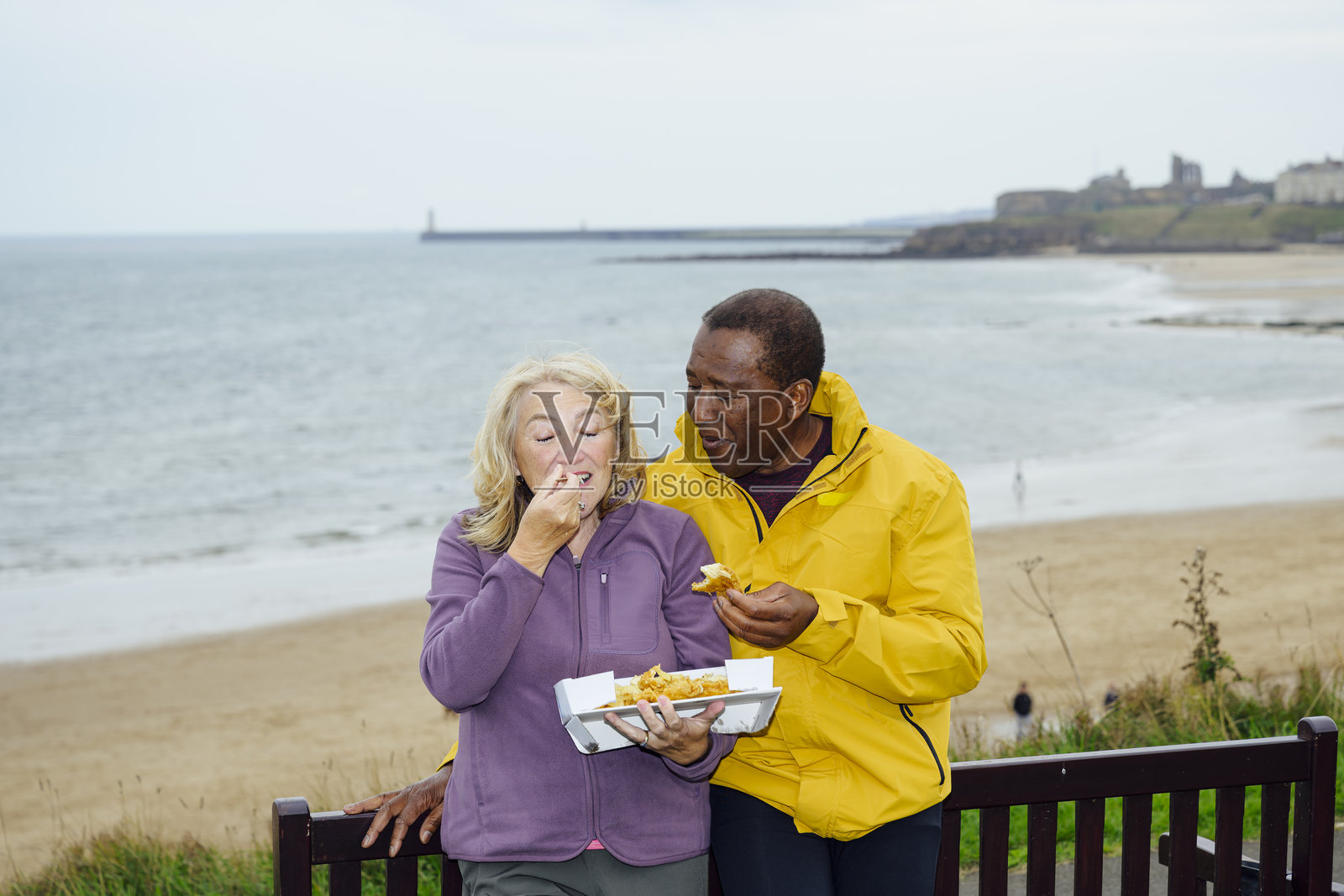 海边背景下，一对年长夫妇正在吃装着炸鱼薯条的盒子照片摄影图片