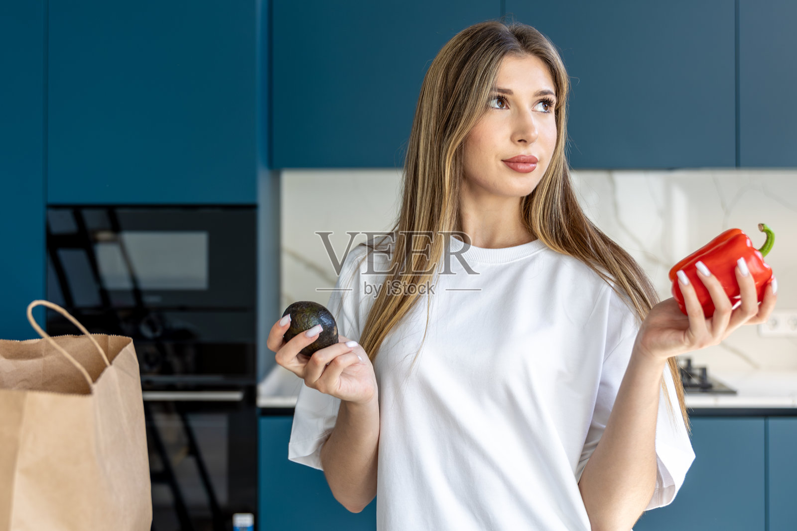 一位女士在厨房里比较牛油果和红甜椒。健康的选择。照片摄影图片