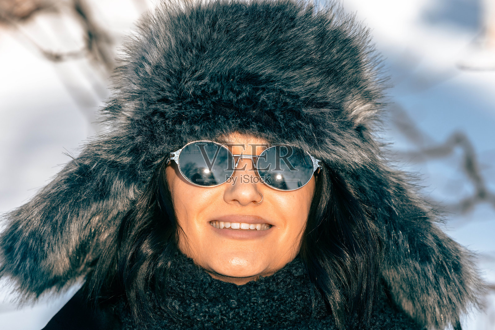 戴着太阳镜和毛绒内衬冬帽的微笑女子，站在雪地户外，在明媚的阳光下展现愉快的冬季体验照片摄影图片