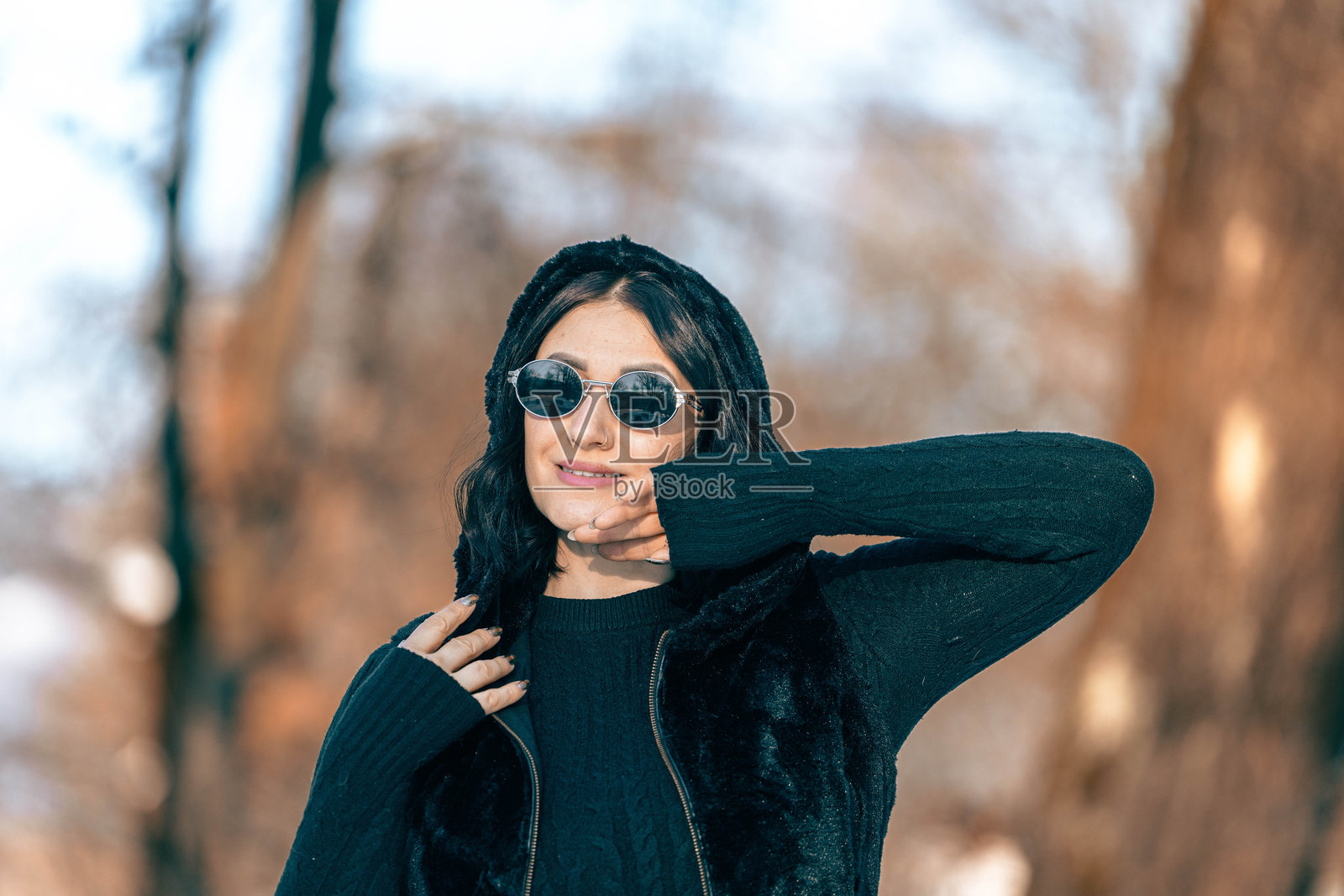 一位留着深色长发的年轻女子戴着太阳镜，穿着黑色毛衣，自信地在户外摆出造型，周围是树木，展现出时尚现代的冬季时尚造型。照片摄影图片