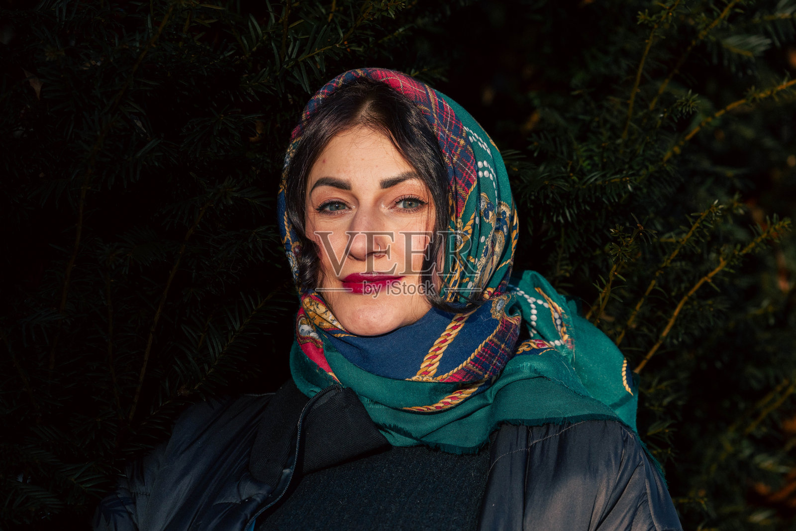 一位深色头发的年轻女子，身着色彩鲜艳的围巾和外套，自信地站在常绿植物背景前，展现出文化时尚与自然之美。照片摄影图片