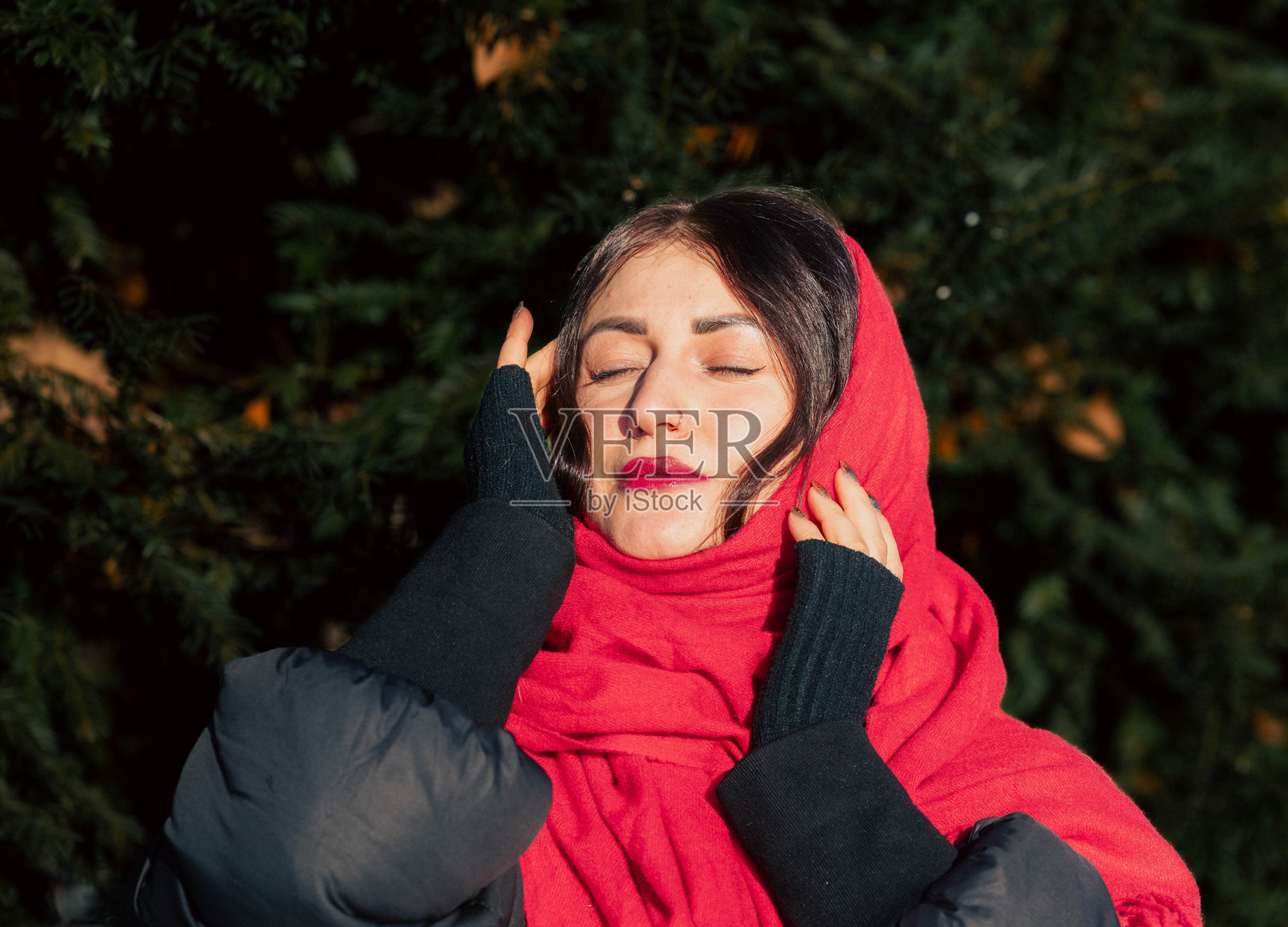 一位留着深色长发的女子，戴着红色围巾和黑色手套，在户外享受阳光明媚的一天，周围环绕着郁郁葱葱的绿树，展现出大自然的温暖与宁静。照片摄影图片