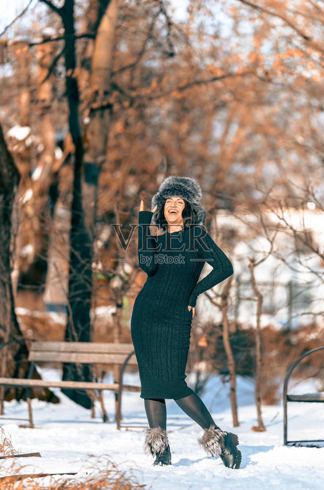 身着黑色针织连衣裙和皮草帽子的女子在雪地公园里快乐地摆姿势，周围有树木和长椅，捕捉到了冬日时尚和幸福的精髓照片摄影图片