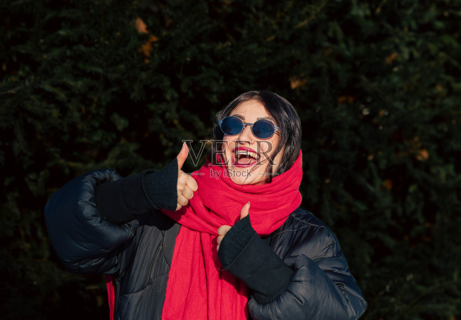 一位留着深色头发、戴着太阳镜、围着红色围巾的年轻女子在户外，周围是茂密的绿色植物，她开心地竖起大拇指，展现出幸福和积极的态度。照片摄影图片