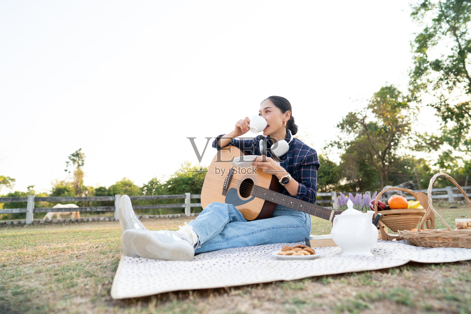 夕阳时分，一位快乐的年轻亚洲女性在乡间公园的野餐垫上放松，喝着咖啡，戴着耳机听音乐，旁边放着一把原声吉他。照片摄影图片
