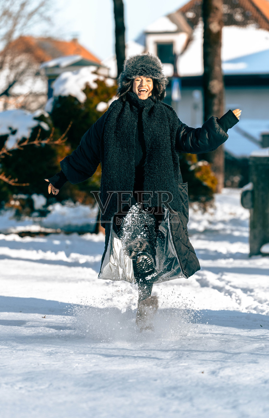 身着冬装的快乐女子在雪地里嬉戏奔跑，周围是树木和温馨的社区，捕捉到了冬季的乐趣和幸福感照片摄影图片