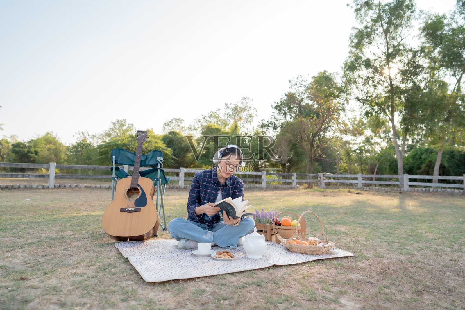 年轻的亚洲女子坐在公园的野餐垫上，戴着耳机，手边放着一把木吉他和茶，正在阅读一本书，享受着悠闲的夏日周末。照片摄影图片