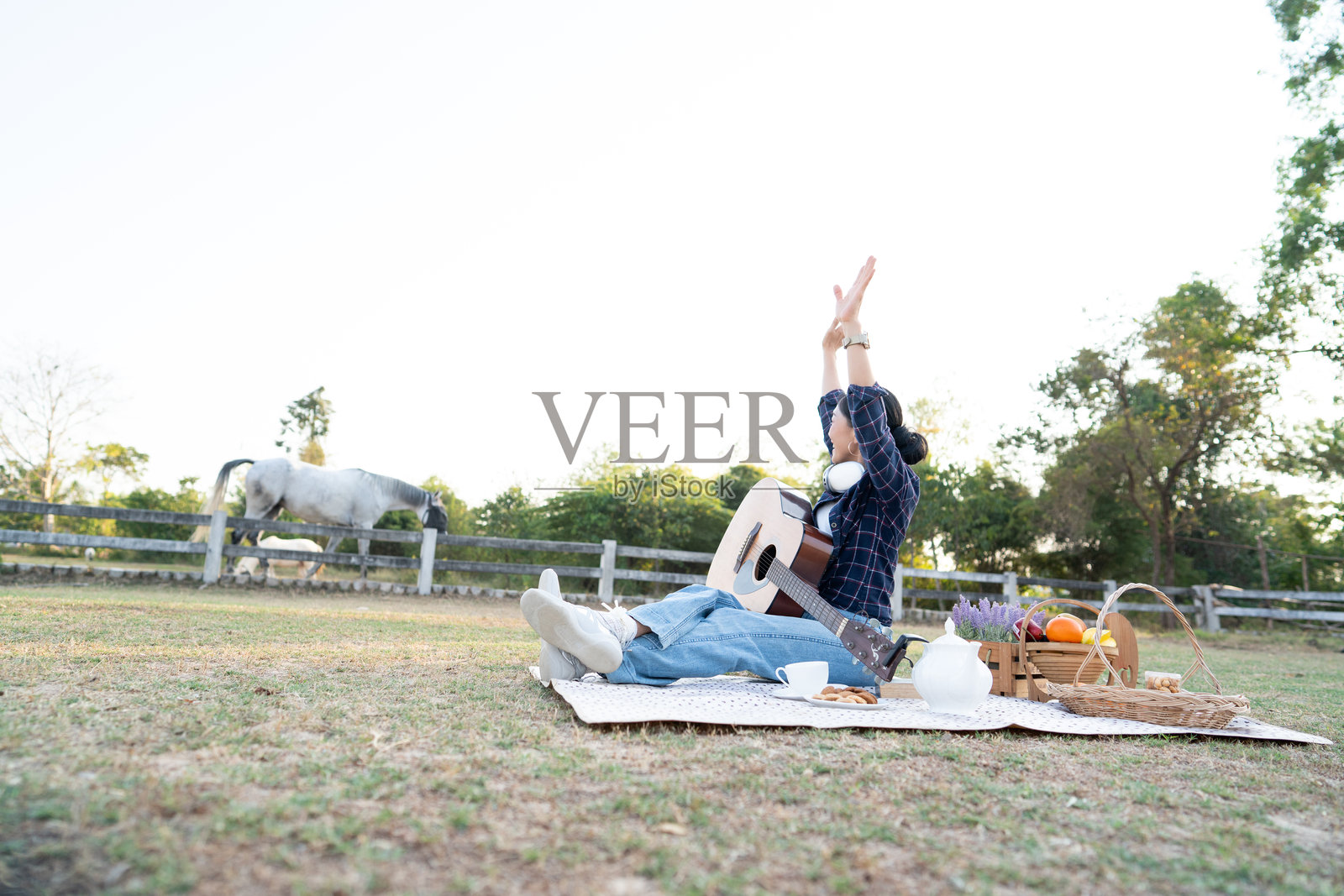 夕阳下，一位快乐的亚洲女性在公园的野餐垫上弹奏原声吉他放松，体现生活方式与音乐爱好概念照片摄影图片