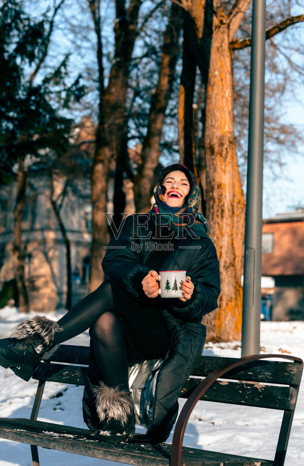 一位身着冬装的女士微笑着坐在公园长椅上，手中拿着一杯热饮，周围是白雪覆盖的树木，在宁静的冬日景观中享受阳光明媚的户外时光。照片摄影图片