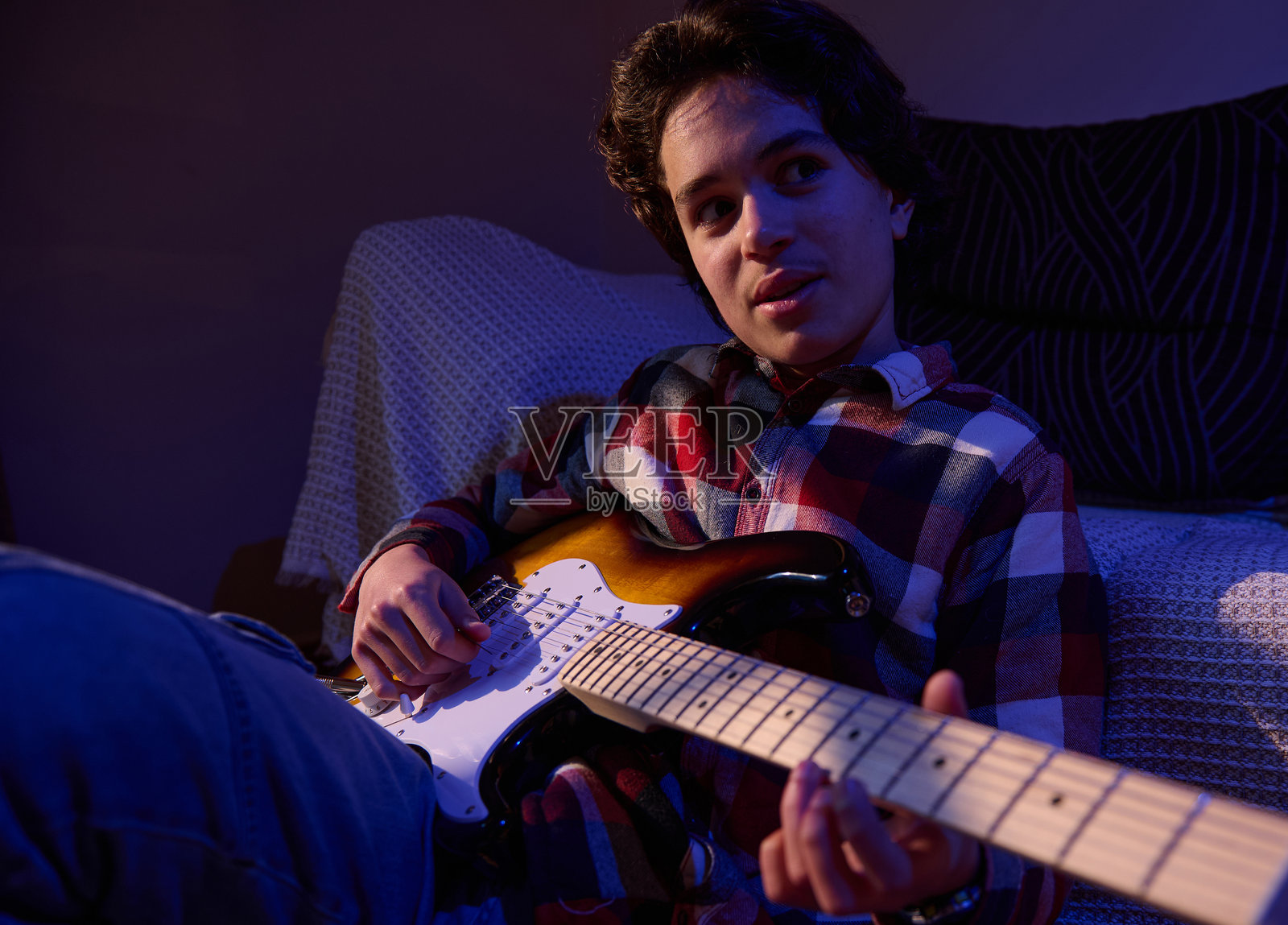 穿着格子衬衫的年轻人在光线昏暗的房间里弹奏电吉他，进行随意的居家音乐练习照片摄影图片