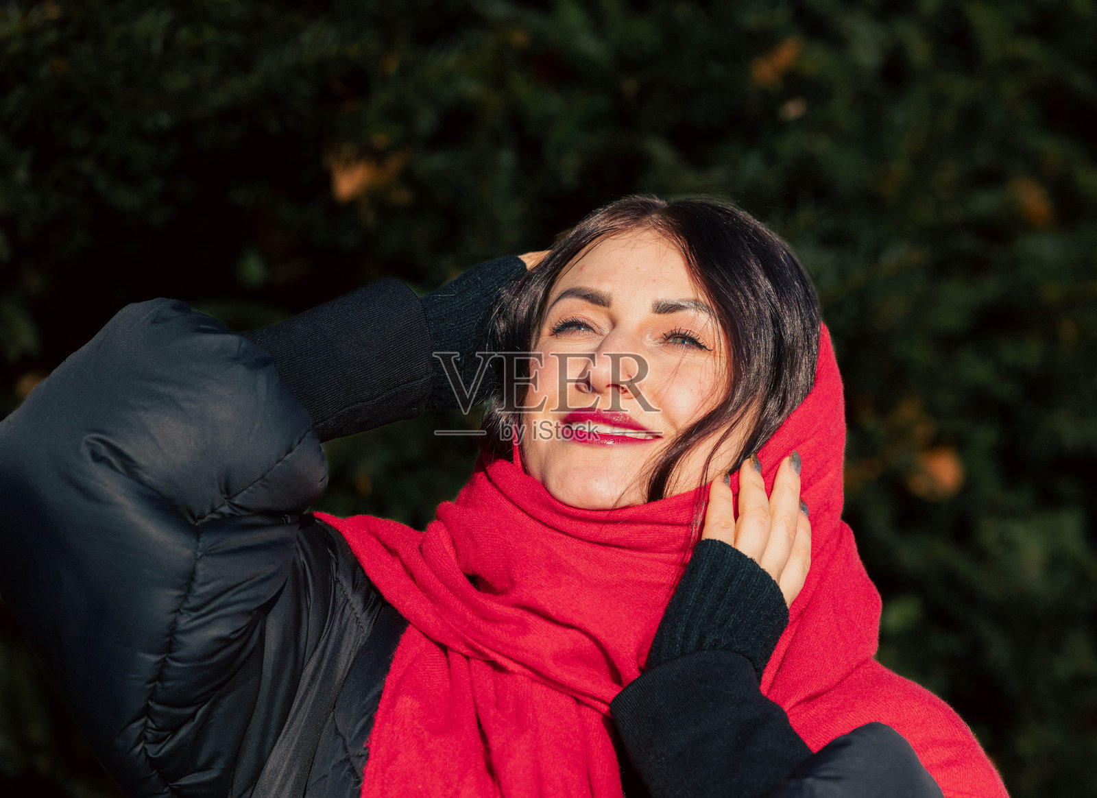 留着深色长发的年轻女子，身穿红色围巾和黑色夹克，在绿意环绕的户外喜悦地微笑，捕捉到了冬日温暖与幸福的精髓照片摄影图片