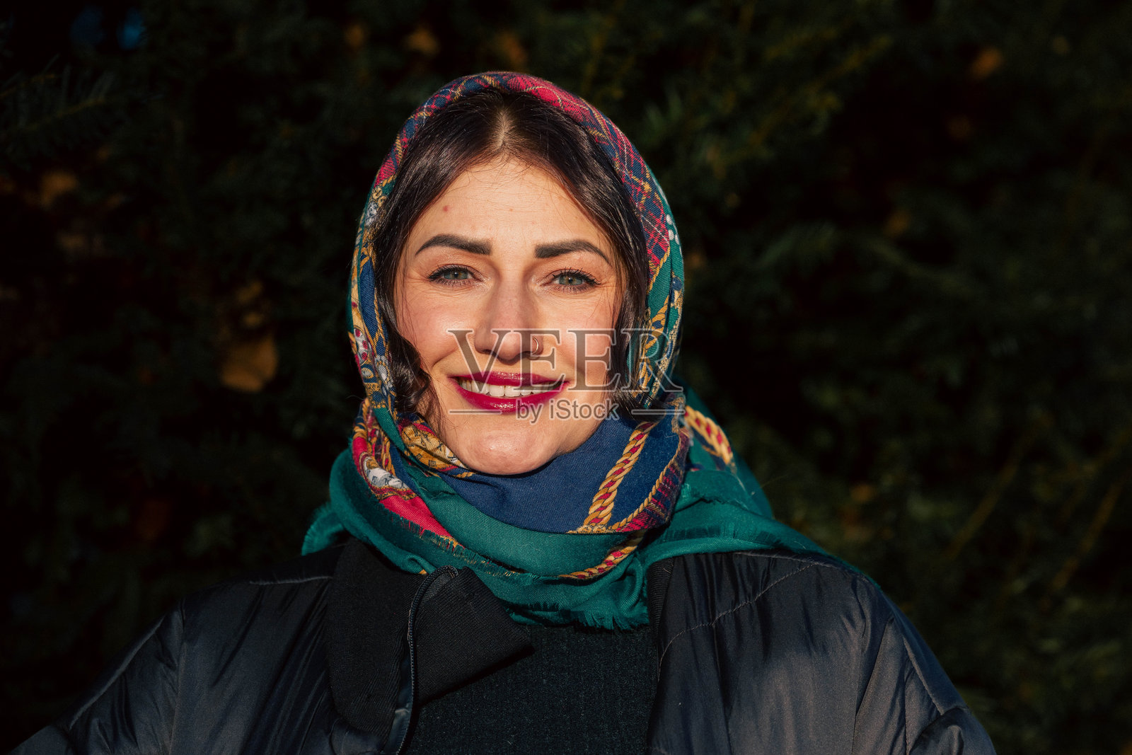 一位戴着彩色围巾、身穿深色外套的微笑女性，站在户外绿植环绕的自然环境中，散发出温暖与喜悦。照片摄影图片